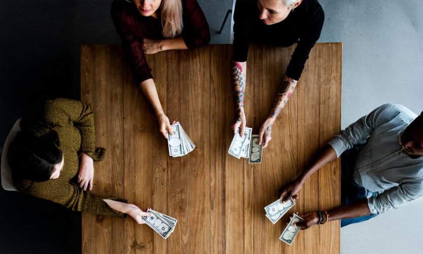 Overhead view of four diverse people sitting around a wooden table, each holding US dollar bills in their hands