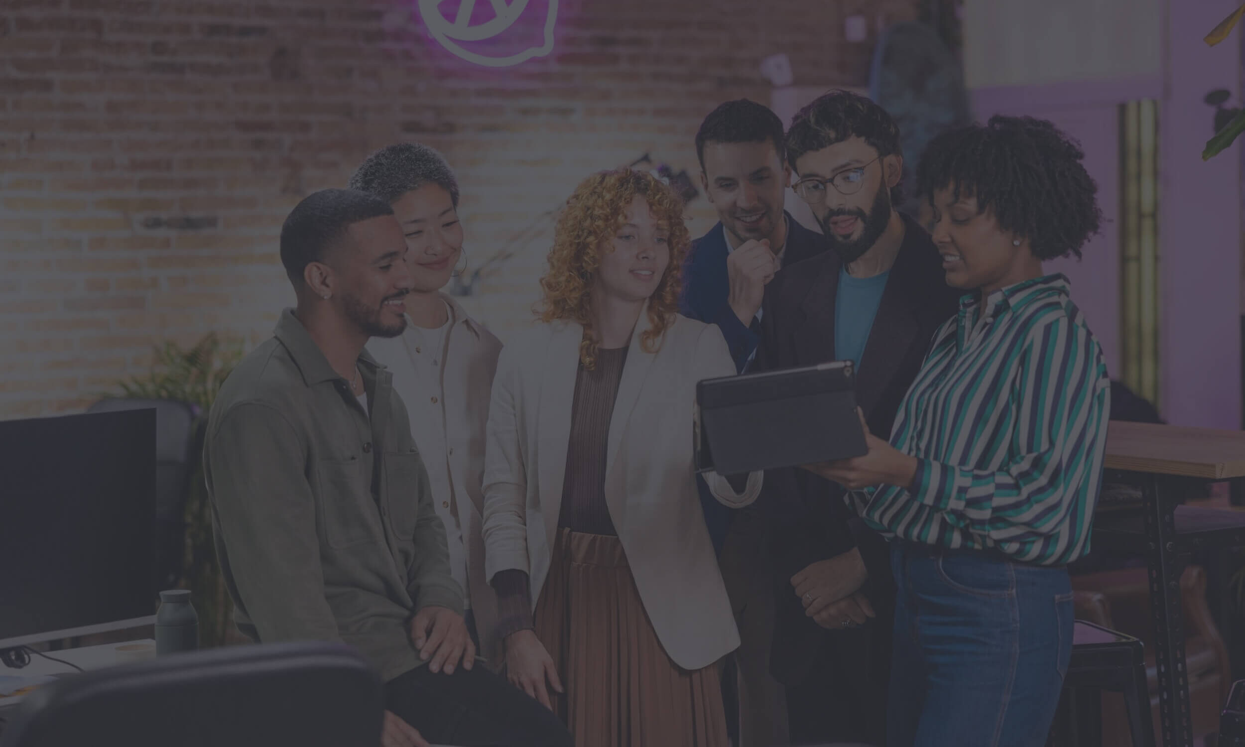 Team of five diverse professionals gathered around a laptop in a modern office, smiling and collaborating while reviewing content on screen