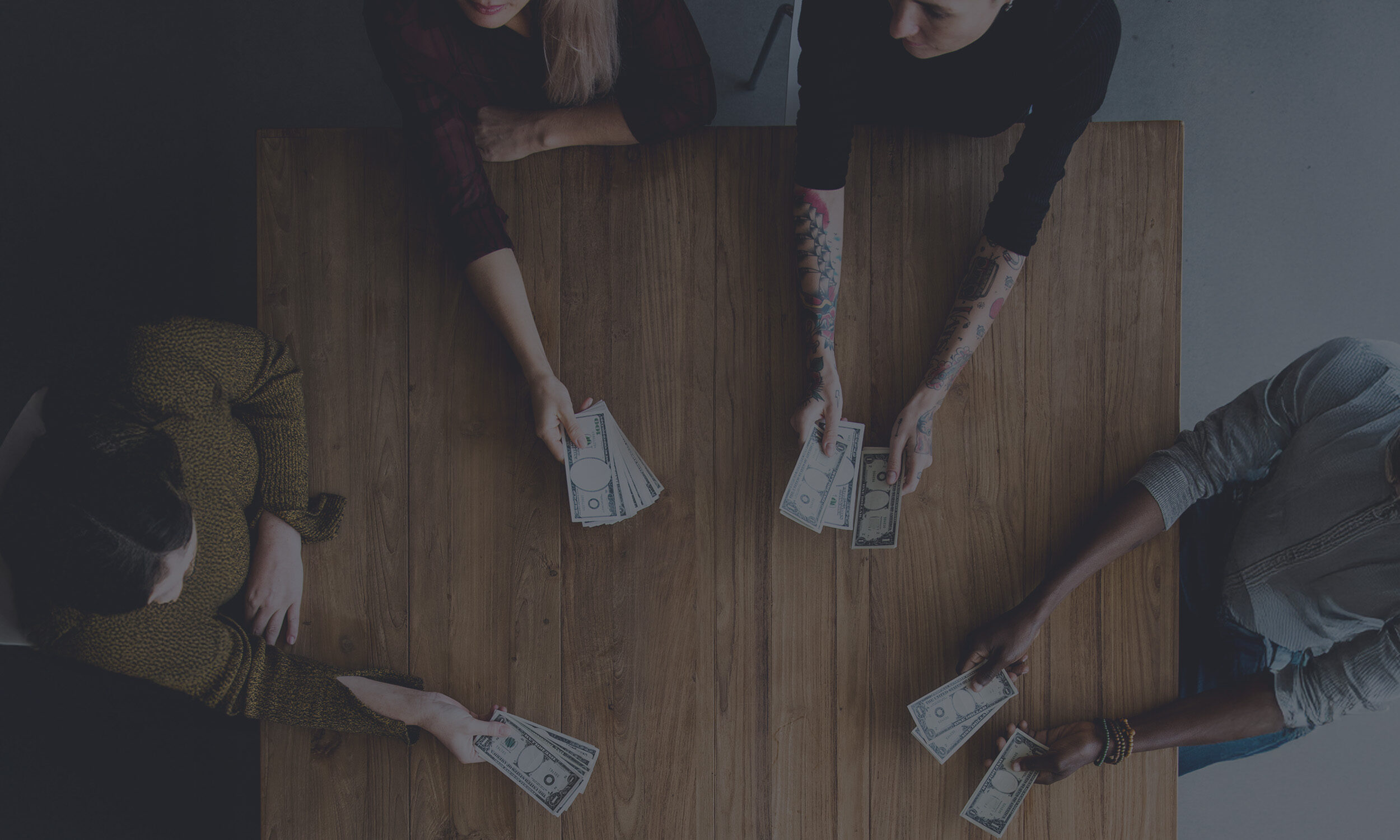 Overhead view of four diverse people sitting around a wooden table, each holding US dollar bills in their hands with a tint overlay