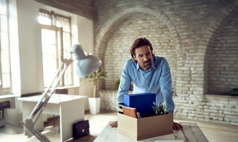 Professional man in blue shirt carrying cardboard box with laptop and belongings, leaving office environment