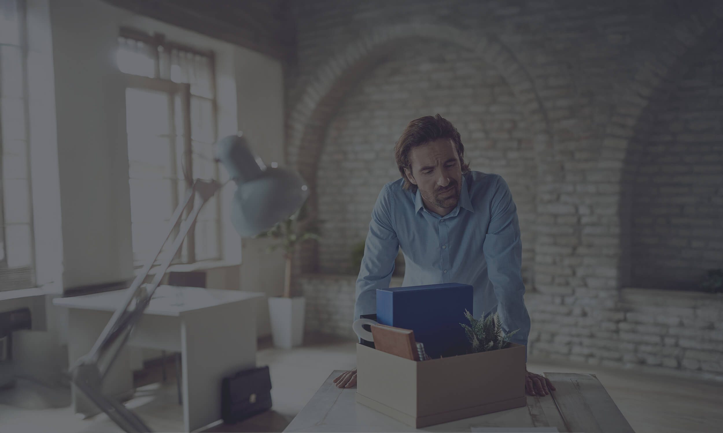 Professional man in blue shirt carrying cardboard box with laptop and belongings, leaving office environment with dark tint.