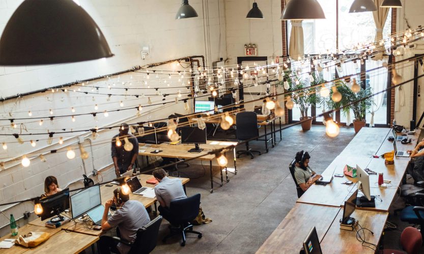 A wide-angle overhead shot of a modern, industrial-style open-plan office. Multiple people are seated at wooden desks working on laptops and desktop computers. Edison bulb string lights hang in rows from the ceiling, giving the space a warm tone beneath the dark grey overlay. Natural light filters in from large windows on the right-hand side. Plants and scattered equipment are visible in the background.