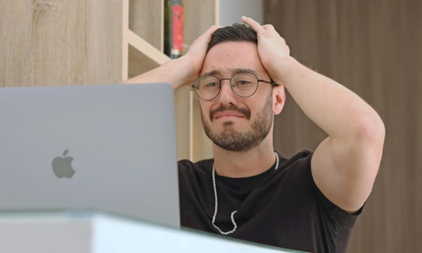 Man sitting at an Apple desktop computer with both hands gripping his hair in frustration, wearing round glasses and a chain necklace