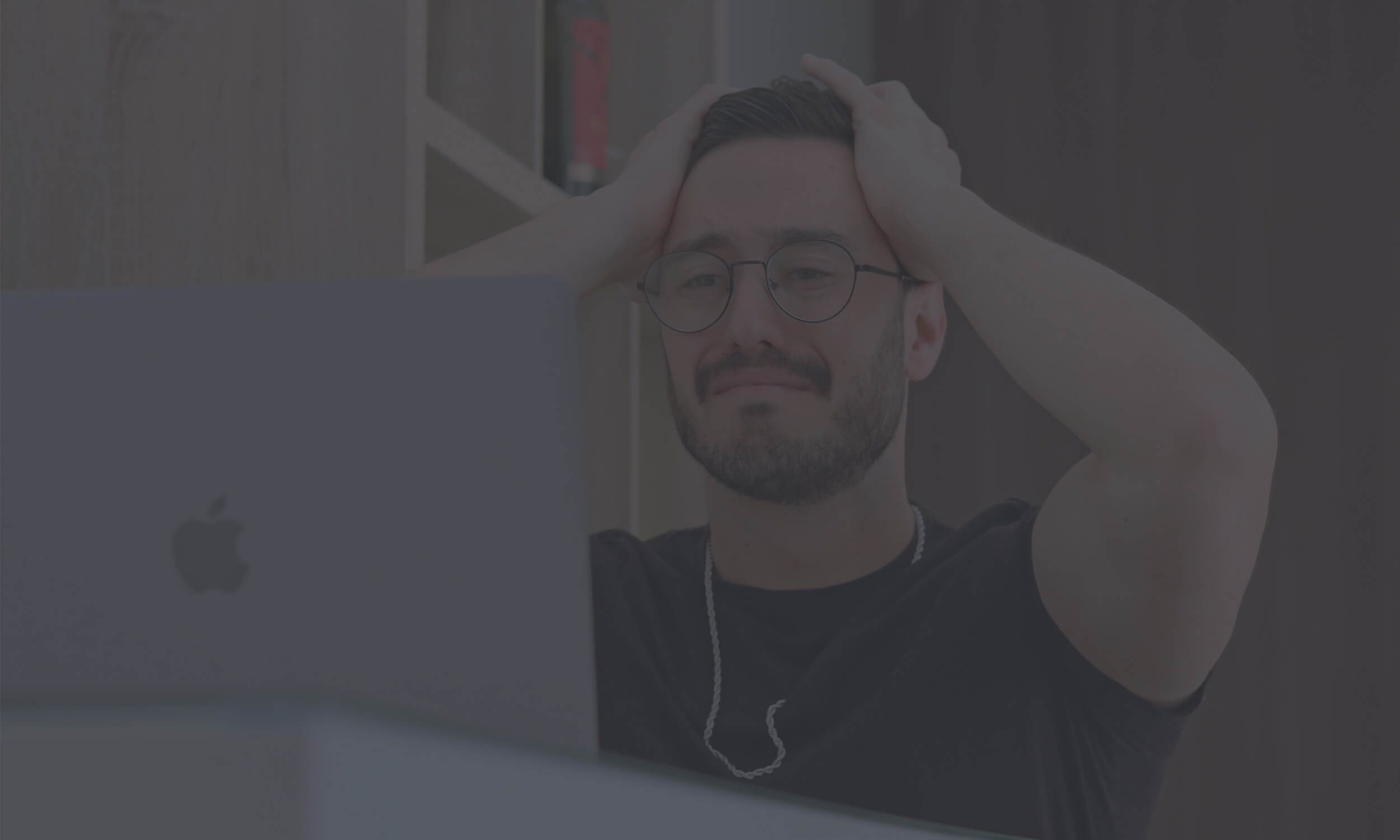 Man sitting at an Apple desktop computer with both hands gripping his hair in frustration, wearing round glasses and a chain necklace